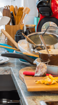 Cluttered kitchen bench stacked with dishes, utensils and food prep mess - typical example of a hoarding affected space before professional decluttering